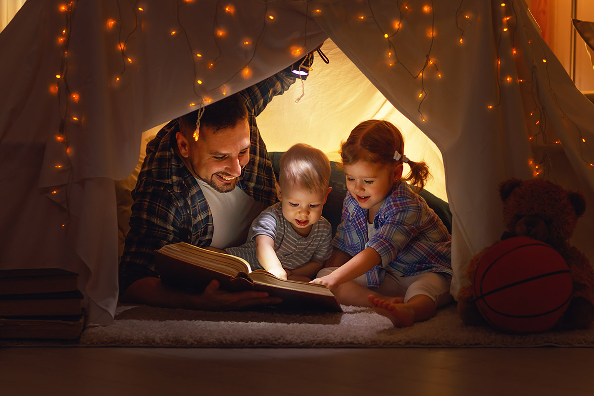 A father sitting in a tent built at home reading to two small children.
