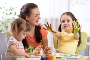 A mother finger painting with two daughters with one daughter holding out her paint-covered hands.