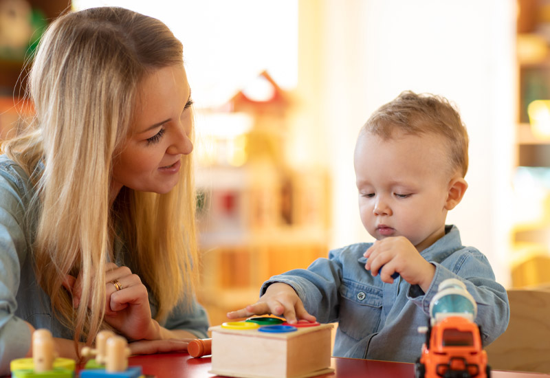 A mom sitting at a table with a small toddler who is playing with wood blocks.