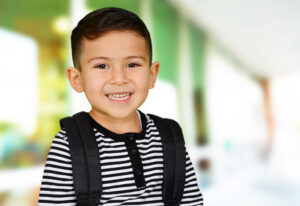 A smiling preschool boy wearing a backpack and a black and white striped shirt.