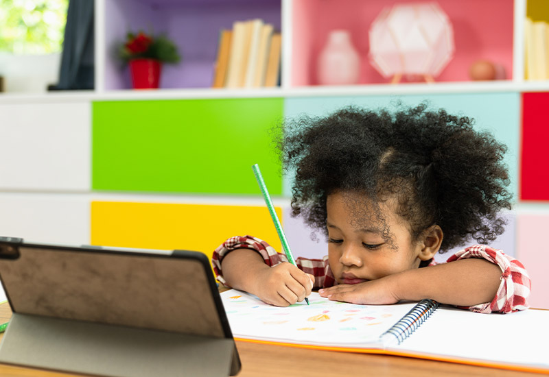 A preschooler sitting at a desk drawing in a notebook with a smart tablet in front of her.